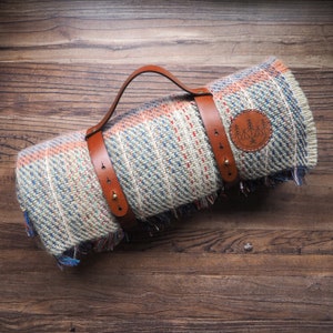 Puede incluir: Manta de picnic enrollada con un patrón tejido en azul, naranja y crema. Se asegura con correas y un asa de cuero marrón. Se ve un parche circular con un diseño de árboles.