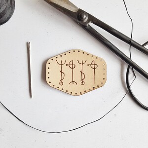 Puede incluir: Primer plano de un proyecto de artesanía en cuero con una aguja, hilo y tijeras. La pieza de cuero tiene un diseño de tres símbolos con un borde punteado.