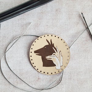 May include: A round, beige leather patch with a brown silhouette of a hand puppet animal and two white hands. The patch is surrounded by a row of small, evenly spaced holes. A needle and thread are visible in the background.