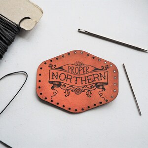May include: A close-up of a leather patch with the text "Proper Northern" in a decorative font. The patch is orange and has holes around the edges for stitching.