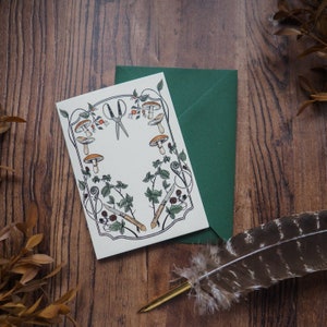 May include: A white card with a black and green border featuring a design of mushrooms, leaves, and small knives. The card is on a wooden surface with a green envelope and a brown feather pen.
