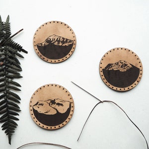 May include: Three brown leather coasters with a black mountain silhouette design. The coasters are arranged in a triangle shape on a white surface. A needle and thread are visible in the background.