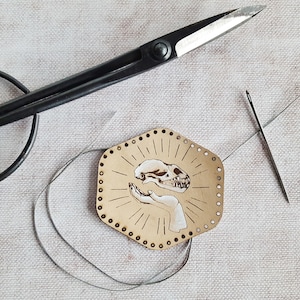 May include: A beige leather craft project featuring a design of a hand holding a skull. The project is being stitched with a needle and thread.