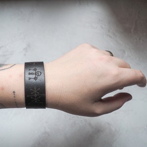 May include: A brown leather bracelet with a geometric design engraved on it. The bracelet is worn on a person's wrist.