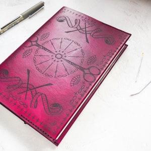 May include: A burgundy leather journal with a decorative design of sewing tools, including scissors, needles, and thread. The design is embossed into the leather and has a vintage aesthetic.