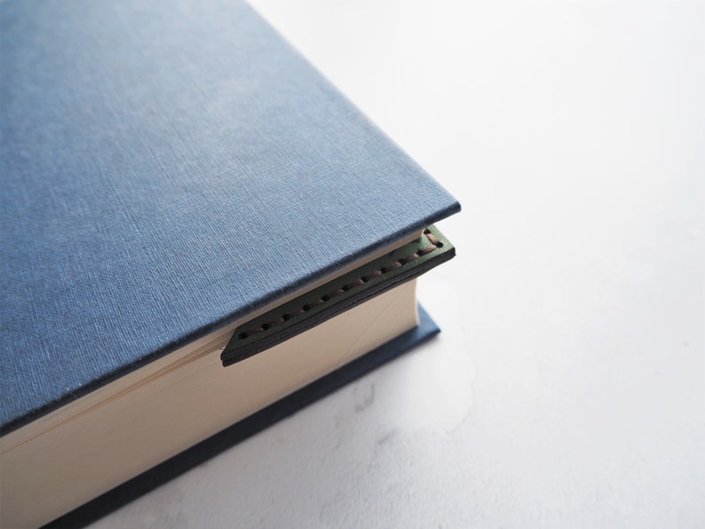 May include: A blue book with a dark green leather bookmark. The bookmark is stitched with red thread and is inserted between the pages. The book has a textured cover and is on a white surface.