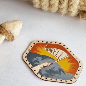 May include: A hand-painted leather patch featuring a mountain scene and the word "SCAFELL" on a banner. The patch has a light tan border with evenly spaced holes. The background has orange and yellow tones.