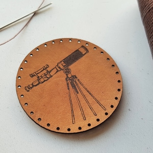 May include: A round, light brown leather piece with a detailed engraving of a telescope on a tripod. The leather has evenly spaced holes around the edge. A spool of brown thread, a needle, and thread are also visible.