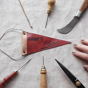 Puede incluir: Un banderín de cuero rojo con el texto "DON'T Panic" en letras negras. El banderín está colgado de una cuerda blanca y negra. Hay varias herramientas de trabajo del cuero alrededor del banderín, incluyendo un pequeño punzón, un cuchillo de cuero, un par de tijeras y un pincel.