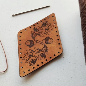 Puede incluir: Un parche de cuero marrón en forma de diamante con un diseño de bellota y hojas. El parche tiene agujeros alrededor de los bordes para coser.