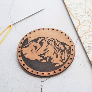 Może przedstawiać: Okrągły, jasnobrązowy kawałek sk&oacute;ry z motywem g&oacute;rskim. Obraz g&oacute;r jest ciemnobrązowy. Sk&oacute;ra ma szereg małych otwor&oacute;w na krawędzi. Ż&oacute;łta nić i igła znajdują się w lewym g&oacute;rnym rogu. Mapa jest w tle.