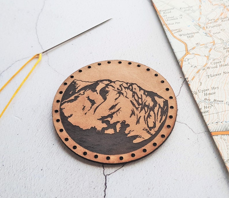 Może przedstawiać: Okrągły, jasnobrązowy kawałek sk&oacute;ry z motywem g&oacute;rskim. Obraz g&oacute;r jest ciemnobrązowy. Sk&oacute;ra ma szereg małych otwor&oacute;w na krawędzi. Ż&oacute;łta nić i igła znajdują się w lewym g&oacute;rnym rogu. Mapa jest w tle.