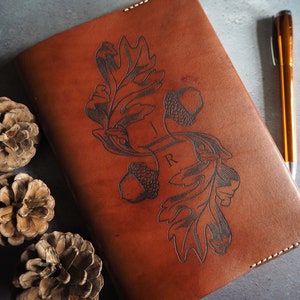 Puede incluir: Un diario de cuero marrón con un diseño decorativo de hojas y bellotas. El diseño está estampado en el cuero e incluye las iniciales "I R".