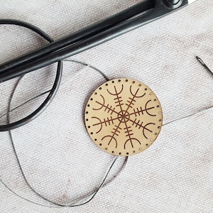 Puede incluir: Un disco redondo de madera con un borde de cuero marrón y un símbolo Vegvisir tallado. Una aguja e hilo están enhebrados a través de los agujeros alrededor del borde del disco. Un par de tijeras negras están sobre una superficie de tela blanca.