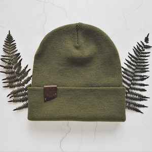 May include: A green knitted beanie with a brown leather patch. The beanie is on a white background with two fern leaves on either side.