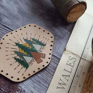 May include: A beige leather patch with a design of three trees and a sun. The trees are green and brown, and the sun is yellow. The patch has a decorative border of small holes.