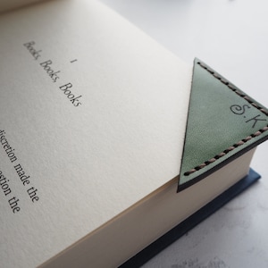 May include: A green leather corner bookmark with brown stitching and the initials "S.K" stamped on it, placed in a book. The book is open to a page with the title "Books, Books, Books".