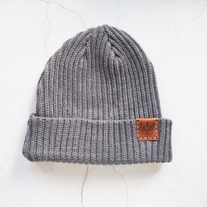 Puede incluir: Gorro de punto gris con un parche de cuero marrón que presenta un diseño de un árbol con un círculo alrededor.