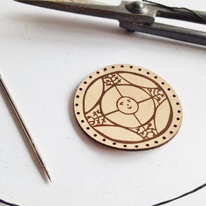 May include: A round, light brown leather craft project with a decorative design of a sun with a face and three stylized flowers. The project is surrounded by a row of small holes.