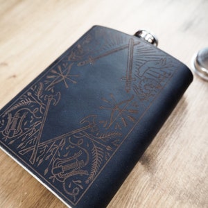 Könnte beinhalten: Eine schwarze Lederflasche mit einem gravierten Design mit Schwertern, Äxten und anderen Waffen. Die Flasche hat einen silbernen Verschluss und einen Trichter.