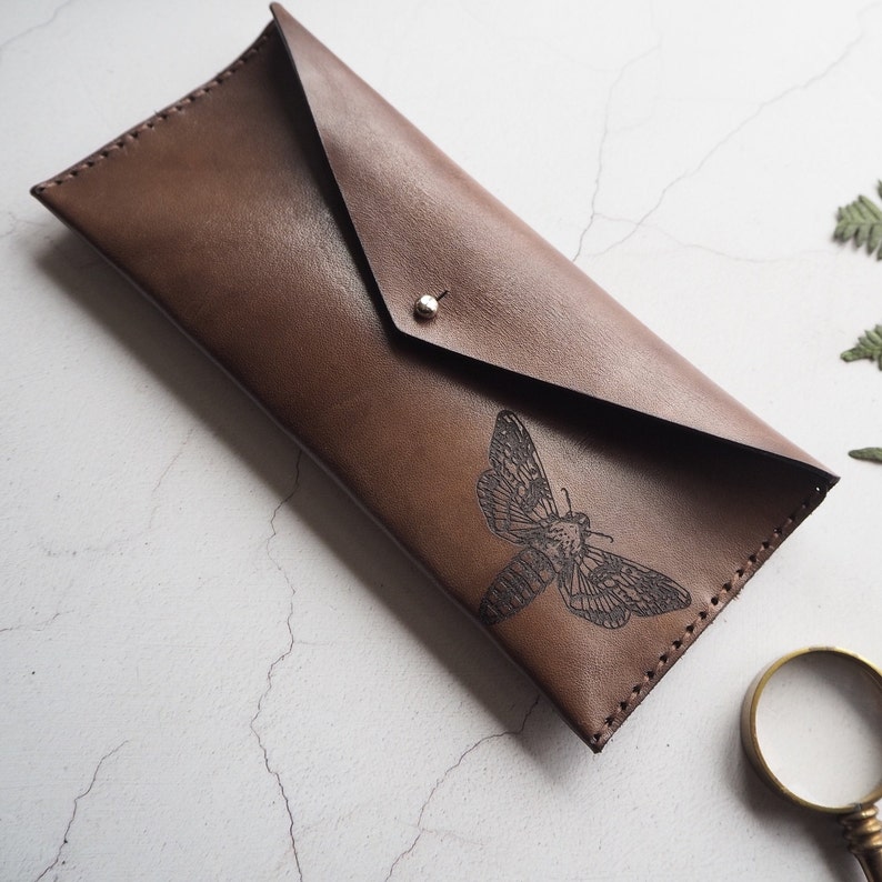 May include: A brown leather envelope clutch with a black moth design. The clutch has a flap closure and a silver rivet.
