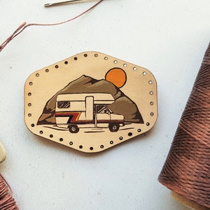 May include: A leather patch with a hand-painted illustration of a camper van driving through a mountain landscape with a sun in the background. The patch has holes around the edge for stitching.