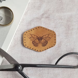 Puede incluir: Un parche de cuero marrón con un diseño de mariposa detallado. El parche tiene una forma hexagonal con un borde cosido.