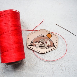 Puede incluir: Una bobina de hilo rojo, una aguja y un parche de cuero con una ilustración de una camioneta vintage, una montaña y un sol. El parche tiene agujeros alrededor del borde para coser.