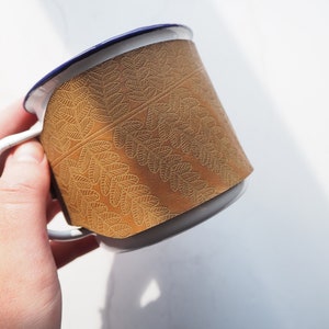 May include: A white enamel mug with a blue rim and a gold leaf patterned band around the middle.