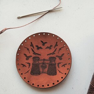 Peut inclure: Un cercle en cuir marron avec des trous autour du bord. Le cercle présente un motif de jumelles et d'oiseaux en vol. Il y a une bobine de fil marron et une aiguille sur une surface blanche.
