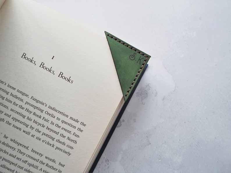 May include: Green leather corner bookmark with stitched edges, resting on the pages of a book. The bookmark has the initials "S.K." embossed on it. The book's title is "Books, Books, Books."