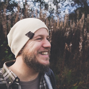 May include: A man with a beard smiles while wearing a white beanie with a brown patch and a plaid shirt.