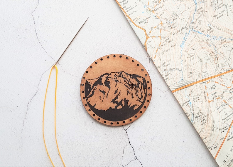 Może przedstawiać: Okrągła, brązowa sk&oacute;rzana naszywka z motywem g&oacute;rskim. Naszywka jest ręcznie szyta wok&oacute;ł krawędzi małymi, r&oacute;wnomiernie rozmieszczonymi otworami. Igła z ż&oacute;łtą nicią i mapa w stylu vintage r&oacute;wnież znajdują się na zdjęciu.