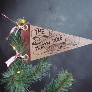 May include: A brown pennant flag with the text "THE NORTH POLE EXPEDITION" in black. The flag is attached to a red leather backing with gold grommets and is decorated with a red and white ribbon. The flag is displayed on a Christmas tree.