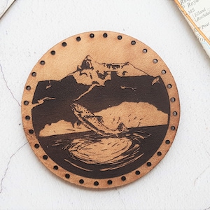 May include: A brown leather coaster with a black and white image of a mountain and a fish jumping out of the water. The coaster has a circle of holes around the edge.