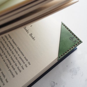 May include: A green leather bookmark with a triangle shape and stitched edges. The bookmark has the initials "S.K." embossed on the front.