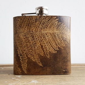 Puede incluir: Una petaca de cuero marrón con una tapa plateada y un diseño de helecho grabado en la parte delantera.