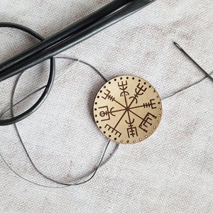 Puede incluir: Una brújula redonda de madera con una aguja negra y un hilo negro. La brújula está rodeada por un círculo de runas. La brújula está sobre una superficie de tela blanca con un par de tijeras negras y una aguja.