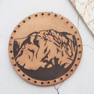 Może przedstawiać: Okrągła, jasnobrązowa sk&oacute;rzana naszywka z ciemnobrązowym wzorem g&oacute;rskim. Naszywka ma szereg małych otwor&oacute;w wok&oacute;ł krawędzi. Mapa i ż&oacute;łta nić w tle.