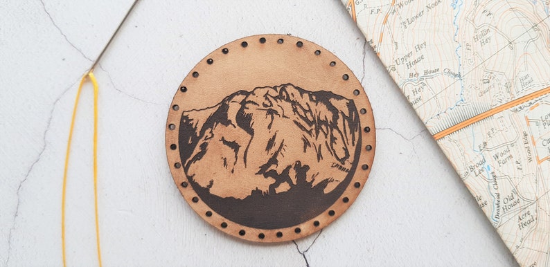 Może przedstawiać: Okrągła, jasnobrązowa sk&oacute;rzana naszywka z ciemnobrązowym wzorem g&oacute;rskim. Naszywka ma szereg małych otwor&oacute;w wok&oacute;ł krawędzi. Mapa i ż&oacute;łta nić w tle.