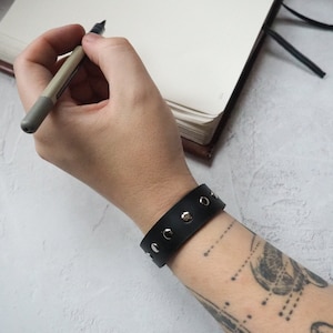 Könnte beinhalten: Ein schwarzes Lederarmband mit silbernen Nieten. Das Armband wird am Handgelenk getragen.