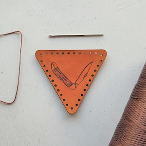 May include: A brown leather triangle with holes punched around the edges and a knife design in the center. A spool of brown thread, a needle, and a length of brown cord are also pictured.