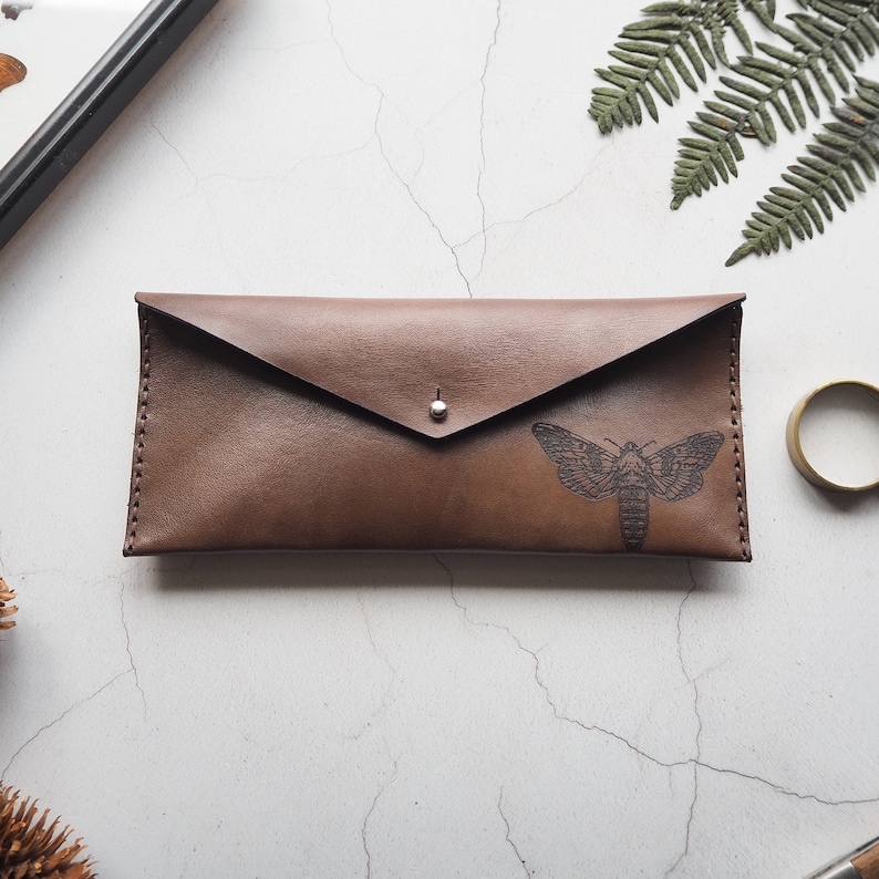 May include: A brown leather envelope clutch with a silver button closure. The clutch has a black moth design embossed on the front.