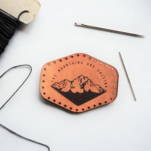 Puede incluir: Un parche de cuero marrón con el texto "THE MOUNTAINS ARE CALLING" y un gráfico de montaña. El parche tiene pequeños agujeros alrededor del borde. Hilo negro y dos agujas también están en la imagen.