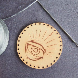 Könnte beinhalten: Ein rundes Stück Leder mit um den Rand herum gestanzten Löchern. Das Leder ist hellbraun und hat ein Design eines Auges mit Lichtstrahlen, die von ihm ausgehen. Eine Nadel liegt neben dem Leder.