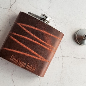 May include: A brown leather-covered flask with a silver top and a funnel. The flask has the words "Courage Juice" engraved on it.