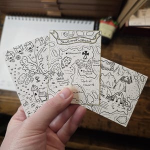 Könnte beinhalten: Drei kleine, handgezeichnete Karten mit schwarzer Tinte auf cremefarbenem Papier. Die mittlere Karte trägt die Aufschrift "Shattered Crescent" und enthält Ortsnamen wie "Glenmere" und "Driftveil Point". Die anderen Karten zeigen Fantasy-Landschaften.