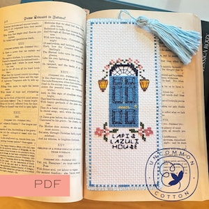 May include: Cross-stitch bookmark featuring a blue door design with the text "LAPIS LAZULI HOUSE." The bookmark has a blue tassel and is placed in an open book with text.