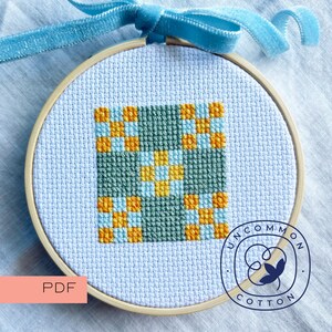 May include: A cross-stitch pattern in a circular wooden embroidery hoop, featuring a geometric grid of dark green, light blue, and yellow squares on white Aida fabric. A light blue velvet ribbon is tied at the top of the hoop for hanging.