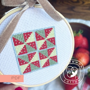 May include: A cross-stitch pattern in a wooden embroidery hoop featuring a geometric quilt block design with red, light green, and dark green triangles on white fabric, accented by a beige ribbon and a PDF label in the corner.